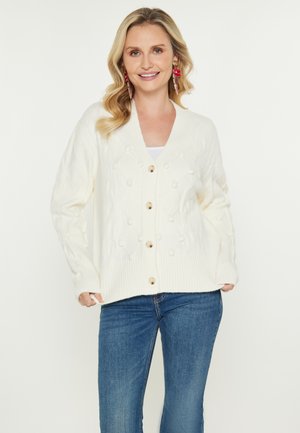Cardigan color crema con scollo a V, caratterizzato da un motivo a diamante, dettagli a pompom e bottoni in legno. Abbinato a jeans blu.