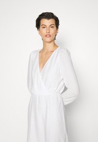 Vestido branco com um padrão texturizado, decote em V e mangas longas. A cintura é acentuada, criando uma silhueta fluida.