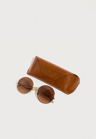 Στρογγυλά καφέ σκουρόχρωμα γυαλιά ηλίου με λεπτά χρυσά πλαίσια δίπλα σε ένα κλειστό καφέ δερμάτινο θήκη πάνω σε λευκό φόντο.