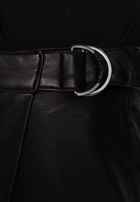 Svart läderbälte med en platt design, som har en rund spänne i silvermetall och en texturerad yta.