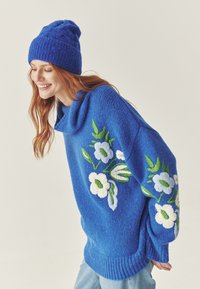 Pulover tricotat bleu cu guler de tip cowl, cu broderie florală în alb și verde. Căciulă tricotată bleu completează ținuta. Blugi bleumarin deschis.
