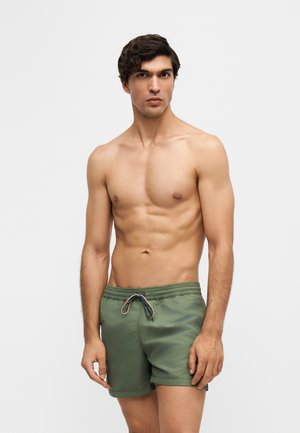 Young man with short curly hair wearing green swim trunks with a drawstring, standing against a plain light background.