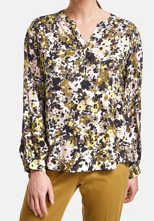 Vrouw die een blouse met lange mouwen en bloemenprint draagt in gele, zwarte en roze tinten, gecombineerd met mosterdgele broek, staande tegen een witte achtergrond.