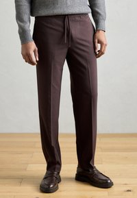 Calças bordô com textura suave, cintura com cordão elástico e corte reto, combinadas com mocassins castanhos num chão de madeira.