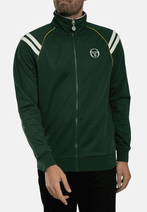 Uomo che indossa una giacca verde scuro con zip, con strisce bianche sulle spalle e un piccolo logo rotondo sul petto sinistro, in piedi con le braccia rilassate.