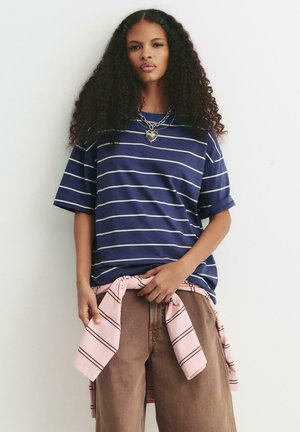 Vrouw met krullend haar, gekleed in een marineblauw gestreept shirt, gelaagde kettingen, een broek in bruine kleur en een roze gestreept shirt om haar middel geknoopt.