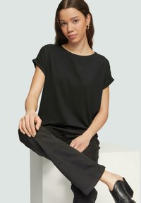 Zwarte korte mouwen t-shirt met opgerolde mouwen, gemaakt van zachte stof. Gedirigeerd met zwarte broeken en hakken, tegen een lichte achtergrond.