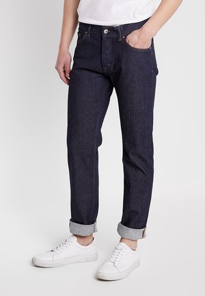Mann trägt dunkle hochgekrempelte Jeans, weiße Sneakers und ein weißes T-Shirt, steht mit einer Hand in der Tasche vor einem einfarbigen Hintergrund.