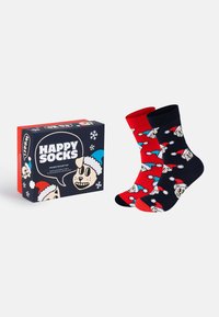 Δύο ζευγάρια κάλτσες με σχέδιο με καπέλα Άγιου Βασίλη και πρόσωπα καρτούν σκύλου, ένα ζευγάρι κόκκινο και ένα ναυτικό, δίπλα σε ένα ταιριαστό κουτί δώρου για τις γιορτές με κατοικίδια.