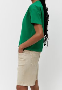Persona con cabello largo trenzado que lleva una camiseta verde de manga corta y pantalones cortos beiges hasta la rodilla, de pie de lado con las manos en los bolsillos.