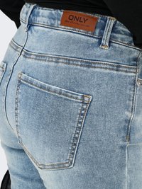 Tuvplāns ar gaisīgi zilām džinsa biksēm no mugurpuses, kabatas vietā ir brūnas ādas etiķete ar uzrakstu "ONLY Blue Denim anno 1995" un oranžu šuvi.