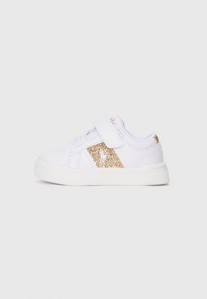 Vit sneaker med glittrande guldaccent på sidan, med en kardborrerem och vit gummisula. Slät material med sömnadsdetaljer.