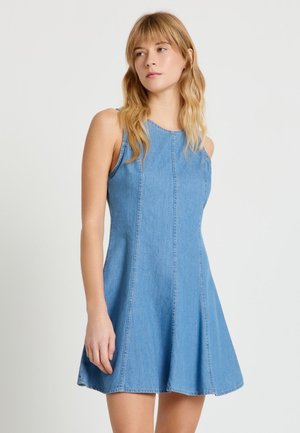 Donna con capelli biondi che indossa un vestito di jeans blu senza maniche, in piedi contro uno sfondo chiaro e uniforme, che guarda di lato.
