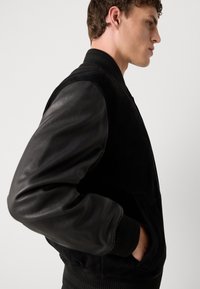 Black bomber jacket featuring suede body and leather sleeves, ribbed cuffs, and a kangaroo pocket, highlighting contrasting textures.