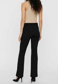 Kvinna med lockigt hår iförd en beige ärmlös topp, svarta högmidjade byxor och svarta högklackade skor, sedd bakifrån.