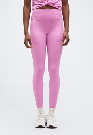 Oseba, oblečena v roza pajkice z visokim pasom, bele nogavice in bele športne copate, stoji ob enobarvnem belem ozadju.