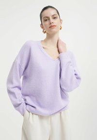 Lavendelfarbener Oversized-Pullover mit V-Ausschnitt, langen Ärmeln und gerippten Bündchen. Weiche Textur und lässige Passform. Kombiniert mit hellen Hosen.