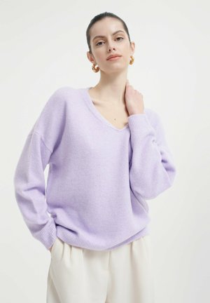 Lila oversized trui met een V-hals, lange mouwen en geribde manchetten. Zachte textuur en een ontspannen pasvorm. Gecombineerd met lichtgekleurde broeken.