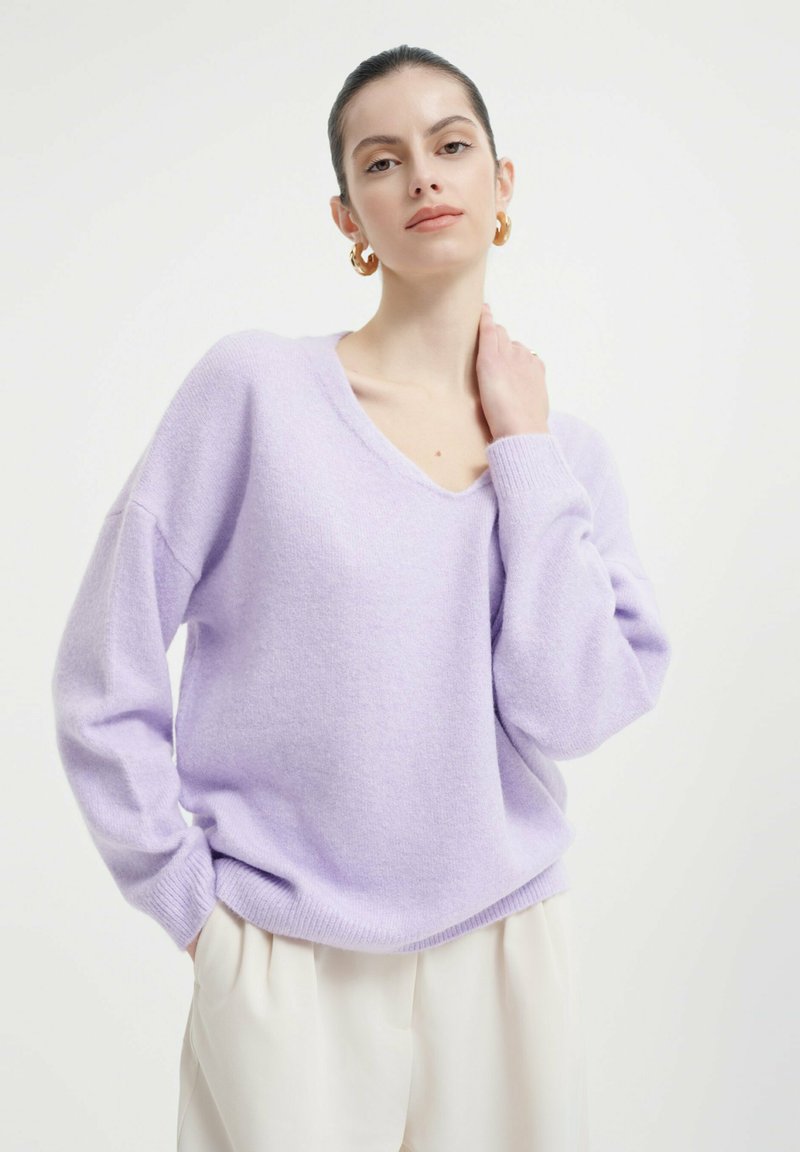 Lavendelfarbener Oversized-Pullover mit V-Ausschnitt, langen Ärmeln und gerippten Bündchen. Weiche Textur und lässige Passform. Kombiniert mit hellen Hosen.