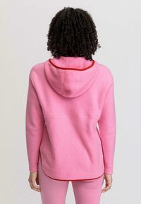 Felpa con cappuccio rosa in pile con un accento rosso attorno al cappuccio, caratterizzata da una vestibilità rilassata e spacchi laterali. Texture liscia, design casual, minimalismo nei dettagli.