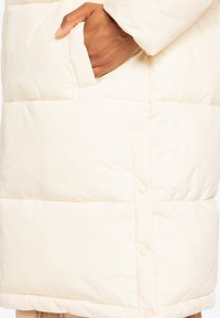 Veste matelassée de couleur crème avec un design en losanges, dotée d'une poche latérale et d'une fermeture à boutons-pression pour une fonctionnalité accrue.
