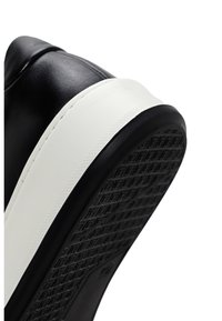 Black leather sneaker with a white midsole and textured black rubber outsole. Features curved design and visible stitching along the edge.