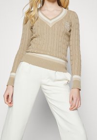 Beige kabelstickad tröja med V-ringning, ribbade ärmslut och nederkant, vita ränder vid halsringning och ärmslut, med en broderad logotyp på bröstet.
