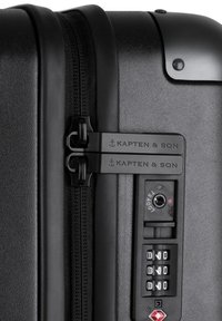 Schwarzer Hartschalenkoffer mit strukturiertem Finish, doppelten Reißverschlüssen, Kapten & Son Branding, integriertem Zahlenkombinationsschloss und TSA-zugelassener Sicherheit.
