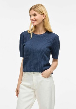Pull en maille bleu marine à manches courtes, avec un motif texturé et des poignets côtelés, associé à un jean blanc.