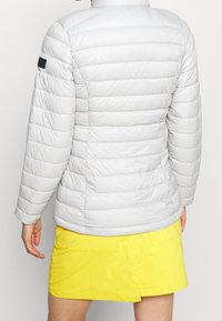 Jasno szara kurtka puchowa z poziomymi pikowanymi panelami, wysokim kołnierzem i czarnym logo na lewym ramieniu. Noszona na żółtej spódnicy.