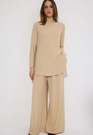 Femme portant une tunique beige à manches longues et un pantalon à jambes larges, debout devant un fond blanc uni, les mains détendues le long du corps.