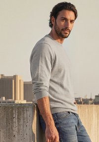 Homme aux cheveux foncés et à la barbe, portant un pull gris clair et un jean bleu, appuyé contre un mur en béton avec des bâtiments de la ville en arrière-plan.