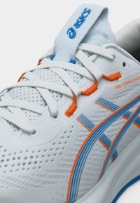 Κοντινό πλάνο λευκού παπουτσιού τρεξίματος Asics GEL-Cumulus 24 με μπλε και πορτοκαλί λωρίδες λογότυπου και λευκά κορδόνια σε αεριζόμενο πλεκτό άνω μέρος.