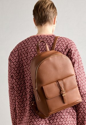 Personne aux cheveux courts portant un pull à motifs et un sac à dos en cuir marron avec une poche avant et une fermeture éclair dorée sur le dos.