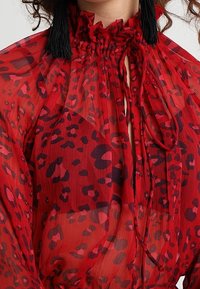 Blouse rouge transparent avec un motif léopard, dotée d'un col montant à volants et de liens, en tissu texturé d'apparence légère et fluide.
