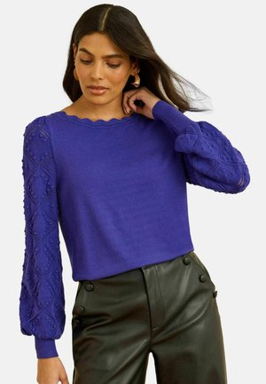Vrouw met lang donker haar, die gouden creolen draagt, een paarse gestructureerde blouse met lange mouwen en een zwarte broek met een hoge taille.