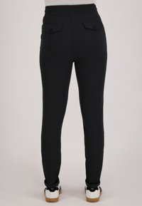 Persoon draagt zwarte strakke broek met knoopsluitingen op de achterzakken en opgerolde pijpen, gecombineerd met witte sneakers met gumzolen.