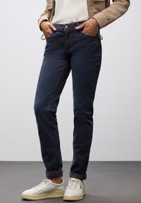 Tmavě modré džíny s úzkým střihem, vybavené knoflíkovým zapínáním, bočními kapsami a ohrnutými manžetami, v kombinaci s bílými teniskami. Hladký povrch.