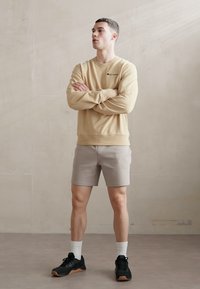 Beige Sweatshirt mit Logo, hellgraue Shorts, schwarze Sportschuhe und weiße Socken; Stehpose vor einem neutralen Hintergrund.