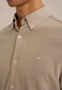 Camisa de botones de color marrón claro hecha de un tejido texturizado, con un cuello estructurado y un pequeño logotipo bordado en el pecho.