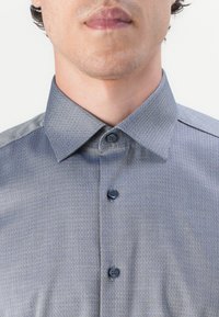 Knappeskjorte i et teksturert mørkeblått stoff med et geometrisk mønster, med spiss krage og to knapper foran.