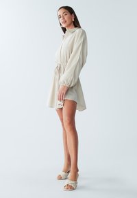 Beige jurk met een kraag en lange mouwen, voorzien van een koord in de taille en een uitlopende zoom. G gedragen met gevlochten beige sliders.