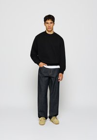 Svart sweatshirt med ribbede mansjetter, kombinert med løse mørke denimjeans og lyse brune sko. Hvit t-skjorte synlig ved kanten.