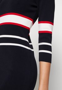 Granatowa dzianinowa sukienka z rękawami o długości 3/4, zdobiona poziomymi paskami w kolorze czerwonym, białym i czerwonym. Gładka tekstura, dopasowany fason.