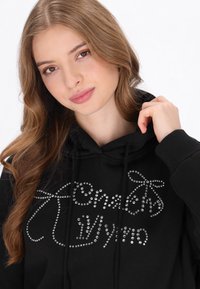 Fekete kapucnis pulóver, strasszkövekkel díszített, írott betűs mintával. A szövet puha, kényelmes szabású, húzózsinóros kapucnival.