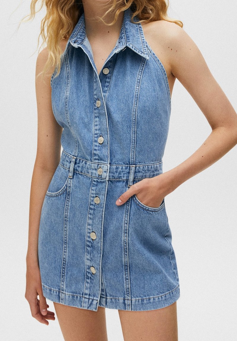 Robe sans manches en denim avec col chemise, boutons sur le devant et deux poches latérales. Tissu bleu clair avec une texture légèrement délavée.