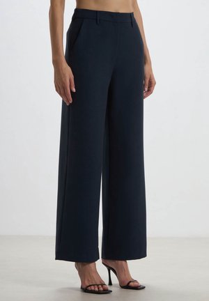 Kobieta nosząca czarne spodnie z szerokimi nogawkami i wysokim stanem oraz czarne sandały na obcasie z odkrytymi palcami, stojąca na jednolitym białym tle.