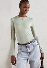 Langermet lysegrønn topp i glatt materiale, sammen med høyde jeans i lyseblå denim og et mørkt belte, med en svart håndveske.
