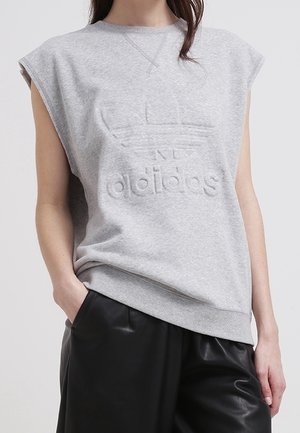Osoba nosząca szarą bezrękawnikową bluzę Adidas z wypukłym logo i czarne skórzane spodnie, ręka w kieszeni, na jednolitym tle.
