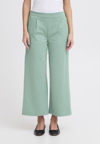 Lichtgroene wijde broek van zachte stof, met een hoge tailleband en plooien voor extra textuur. Gepaarde met zwarte platte schoenen.